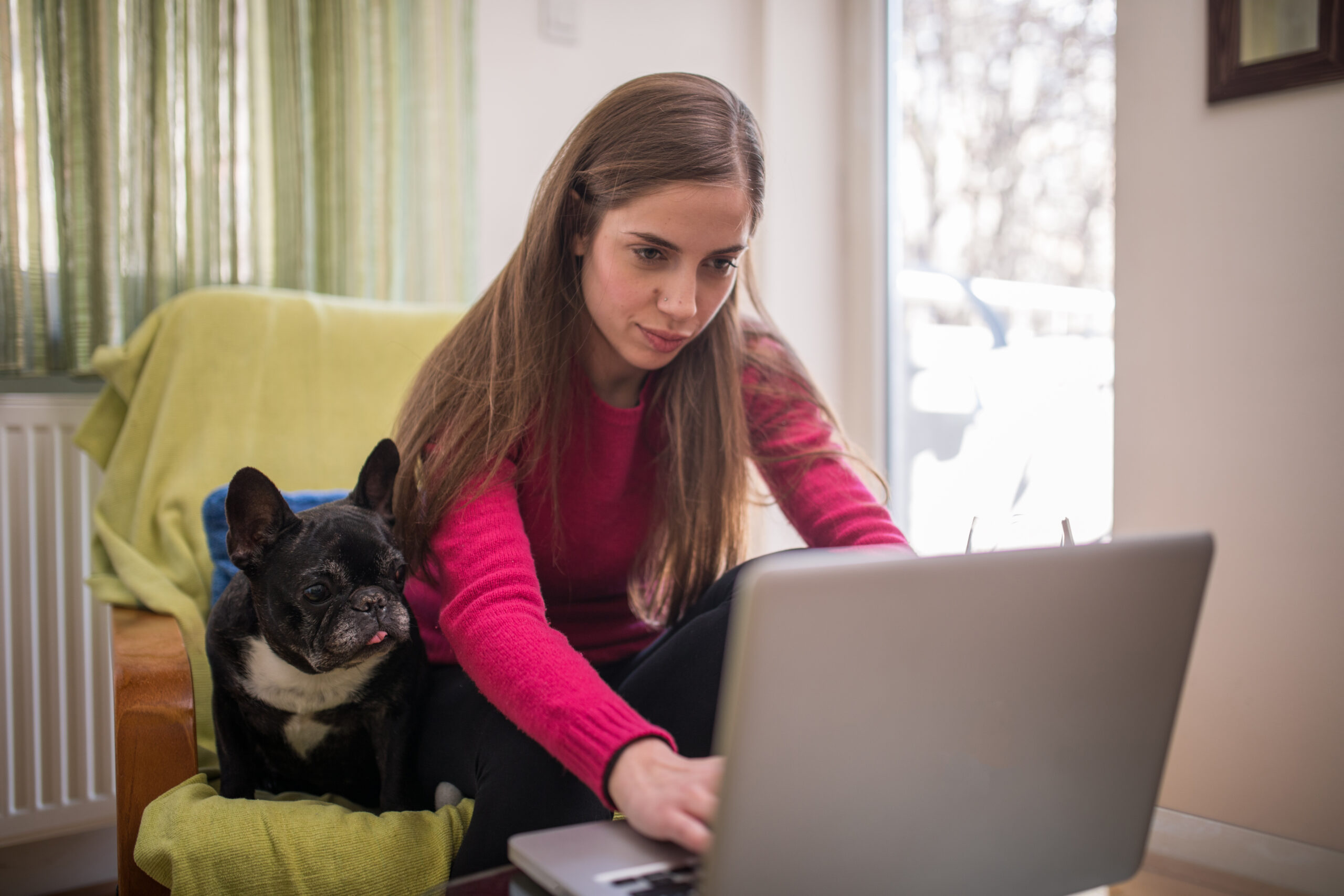 Nainen istuu nojatuolissa ja näpyttää pöydällä olevaa tietokonetta. Koira istuu naisen vieressä ja katsoo myös konetta.